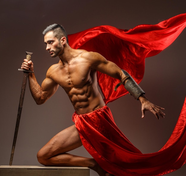 Macho sem camisa em armadura romana com espada em movimento. pano vermelho sobre fundo cinza