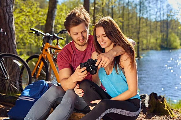 Macho sorridente mostrando fotos de passeios de bicicleta para sua namorada na costa do rio selvagem.