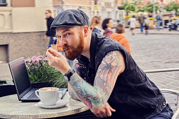 Macho tatuado barbudo vestido com um colete e boné de tweed usando laptop em um café de rua de verão.