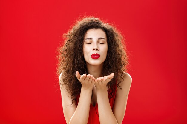 Macia e gentil elegante caucasiana garota com penteado encaracolado e batom vermelho, curvando-se em direção à câmera com um leve sorriso, fechou os olhos e as palmas das mãos perto dos lábios dobrados, mandando um beijo romanticamente para a câmera.