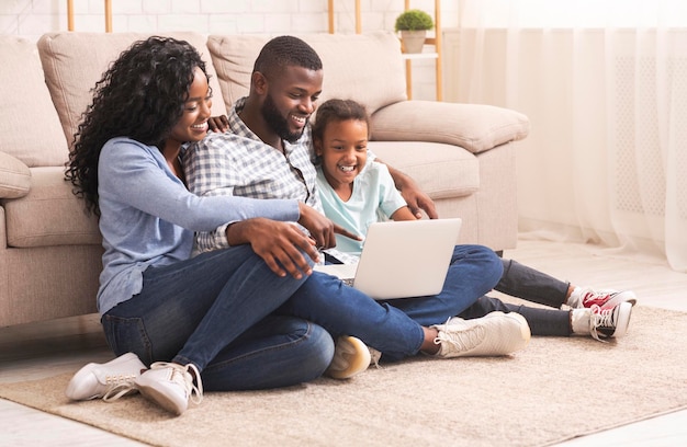 Mãe afro-americana, pai e filha usando laptop juntos assistindo fotos de família enquanto está sentado no chão em casa.