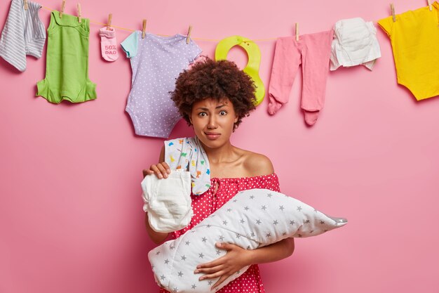 Mãe afro-americana segura a fralda suja e sente aversão, pega um acre de criança que dorme profundamente durante o dia, está ocupada amamentando o recém-nascido, fica encostada na parede rosada, pendurando roupas de bebê lavadas atrás