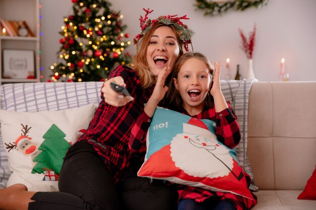 Mãe alegre com coroa de azevinho segurando o controle remoto da TV com a filha sentada no sofá e curtindo o Natal em casa