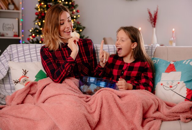 Mãe alegre segura batatas fritas e gesticula para esperar a filha de olhos fechados coberto com um cobertor sentada no sofá e curtindo o natal em casa