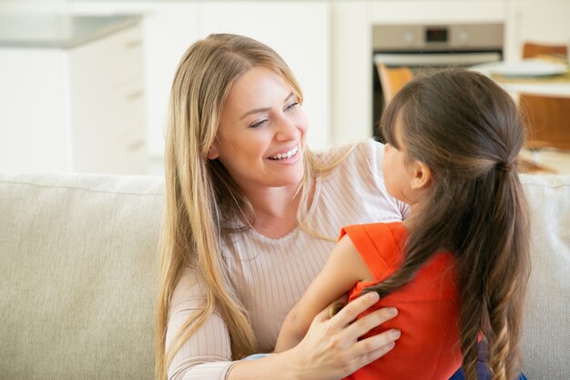 Mãe alegre segurando sua filha nos braços no colo, conversando com ela e rindo.