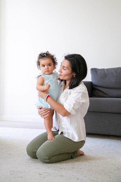 Mãe alegre sentada no chão em casa, segurando a doce filha nos braços. Tiro vertical. Conceito de paternidade e infância