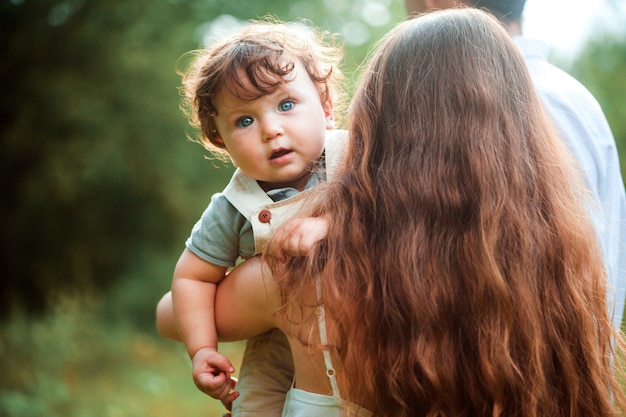 Mãe bonita nova que abraça seu filho pequeno da criança contra a grama verde. Mulher feliz com o filho dela em um dia ensolarado de verão. Família caminhando no Prado.