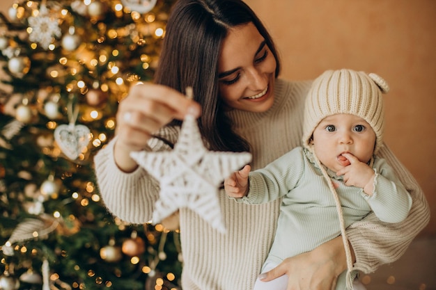 Mãe com a filha segurando um brinquedo de natal perto da árvore de natal