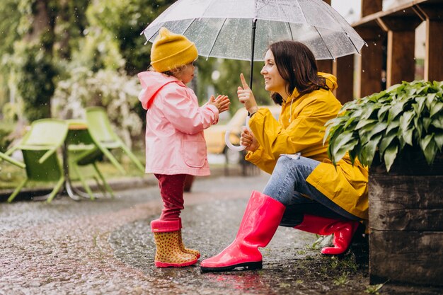 Mãe com filha caminhando no parque na chuva usando botas de borracha