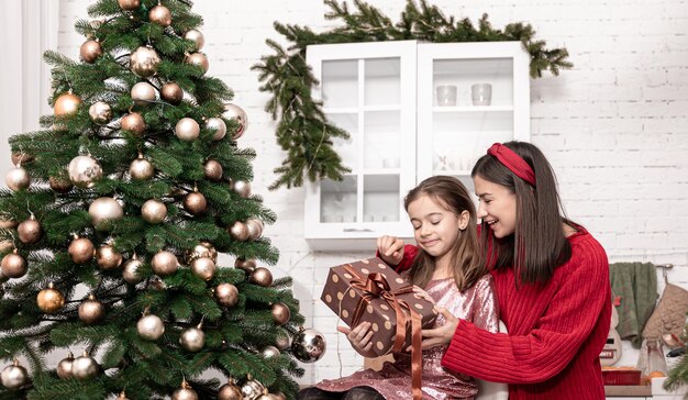 Mãe com uma filha perto da árvore de natal com uma caixa de presente