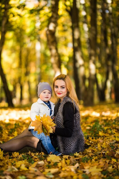 Mãe de família feliz brincando com a criança no parque outono perto de árvore deitada sobre folhas amarelas. Conceito de outono.