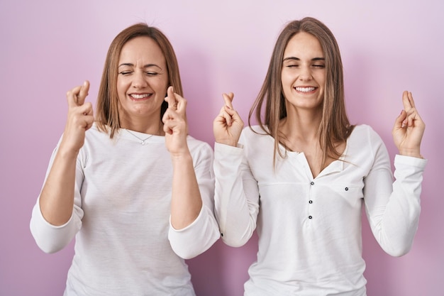 Mãe de meia-idade e filha jovem de pé sobre fundo rosa gestando dedo cruzado sorrindo com esperança e olhos fechados sorte e conceito supersticioso