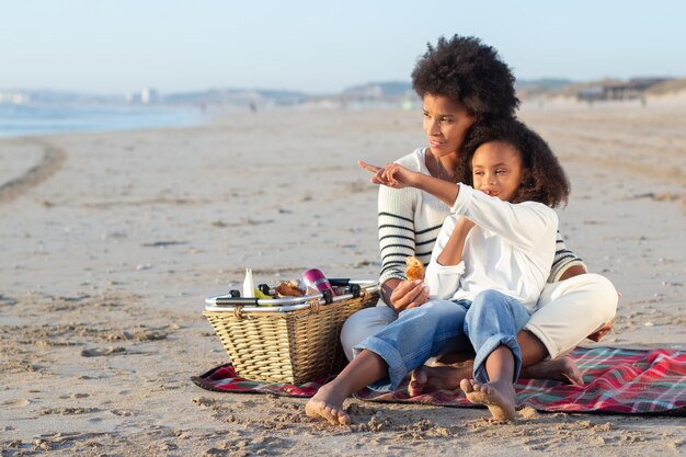 Mãe e filha afro-americanas no piquenique na praia. Mulheres em roupas casuais sentadas no cobertor, comendo pães, olhando para a água. Família, relaxamento, conceito de natureza