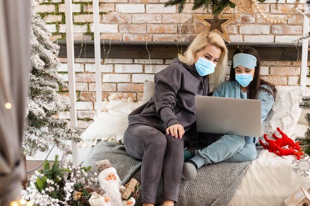 Mãe e filha com máscaras médicas no rosto ligam para parentes em um computador para desejar feliz natal.