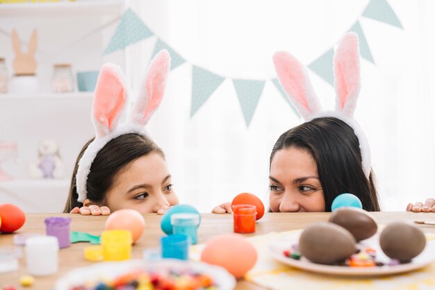 Mãe e filha com orelhas de coelho, espreitando por trás da mesa com saborosos ovos de Páscoa