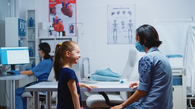 Mãe e filha conversando e esperando o médico no consultório médico durante o coronavírus. Especialista em medicina com máscara de proteção prestando serviços de saúde, consulta, tratamento em hospital.