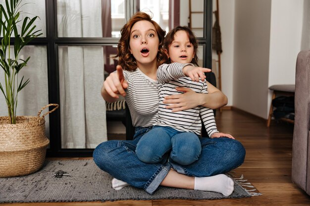 Mãe e filha em jeans idênticos e camisetas estão olhando para a câmera de surpresa e mostrando o dedo