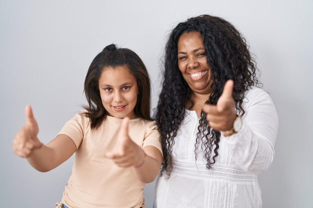 Mãe e filha em pé sobre fundo branco, apontando os dedos para a câmera com cara feliz e engraçada, boa energia e vibrações