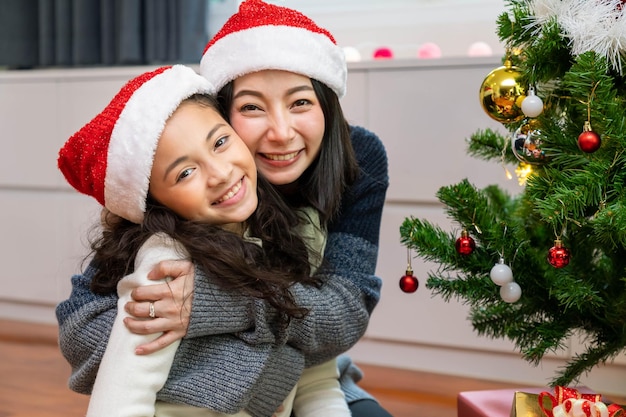 Mãe e filha estão decorando a árvore de Natal juntas para o festival na sala de estar olhando para a câmera