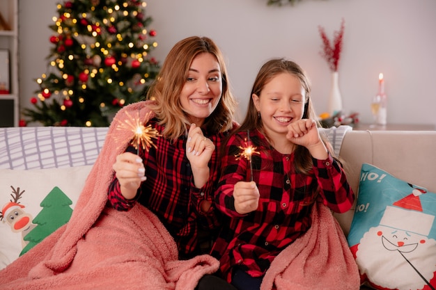 Mãe e filha satisfeitas segurando e olhando para estrelinhas cobertas com cobertor, sentadas no sofá e curtindo o natal em casa