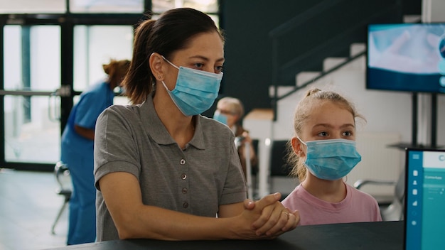 Mãe e filho com máscara conversando com recepcionista no balcão de recepção do hospital, perguntando sobre o formulário de consulta médica. pessoas esperando para comparecer à consulta durante a pandemia de covid 19.