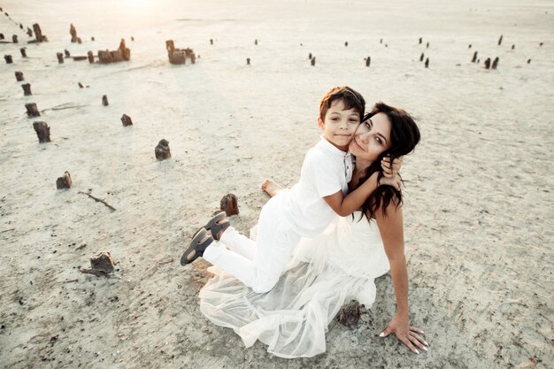 Mãe e filho estão sentados na areia, vestidos com roupas brancas, sorrindo e abraçando