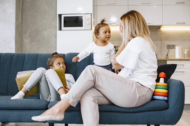 Mãe e filhos relaxando juntos no sofá da sala de estar em casa. meninas brincando com brinquedos e lendo um livro.