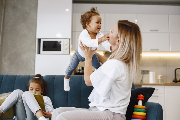 Mãe e filhos relaxando juntos no sofá da sala de estar em casa. Meninas brincando com brinquedos e lendo um livro.