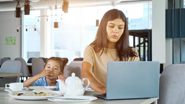 Mãe empresária asiática trabalha sentada à mesa com chá e laptop cinza e criança come fatia de pizza t. Foto Premium