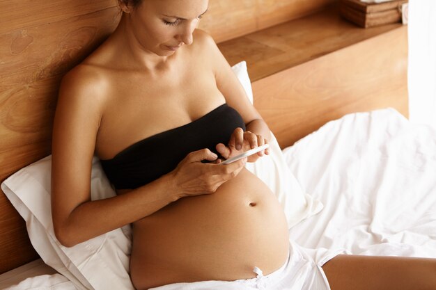 Mãe expectante bonita na parte superior cortada preta que descansa na cama