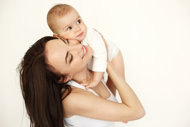 Mãe feliz bonita nova que sorri com os olhos fechados que guardam sua filha do bebê sobre a parede branca.