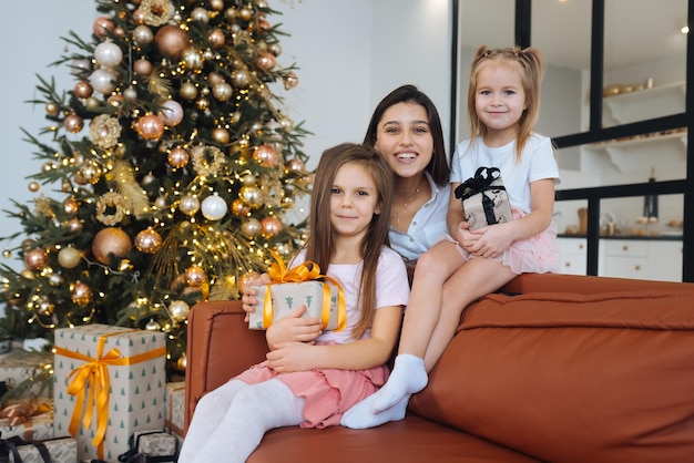 Mãe feliz e duas filhas estão sentadas no sofá e sorrindo contra o fundo da árvore de Natal