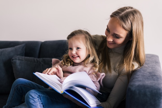 Mãe feliz e filha lendo livro, sentado no sofá confortável na sala de estar