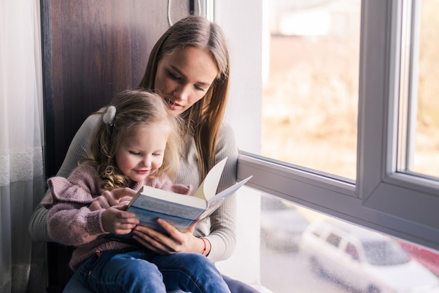 Mãe feliz e filha lendo livro, sentado no sofá confortável na sala de estar