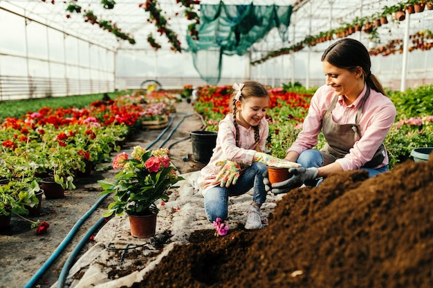 Mãe feliz e sua filha pequena trabalhando com solo enquanto planta flores em uma estufa