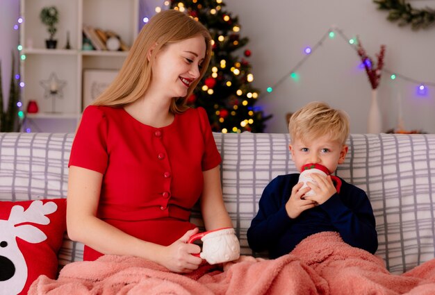 Mãe feliz em um vestido vermelho com seu filho pequeno sentado em um sofá sob o cobertor bebendo chá em canecas em um quarto decorado com árvore de Natal na parede