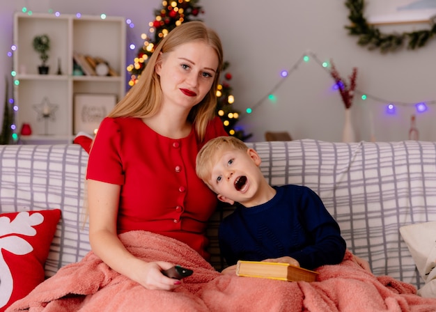 Mãe feliz em um vestido vermelho com seu filho pequeno sob o cobertor lendo um livro em um quarto decorado com uma árvore de Natal na parede