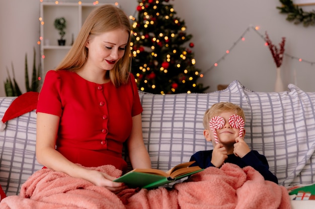 Mãe feliz em um vestido vermelho sentada em um sofá com seu filho pequeno sob o cobertor lendo um livro em um quarto decorado com uma árvore de natal na parede