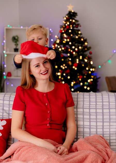 Mãe feliz em um vestido vermelho, sentada em um sofá, sorrindo, enquanto seu filho pequeno está atrás de colocar o chapéu de Papai Noel na cabeça de sua mãe em um quarto decorado com árvore de Natal na parede
