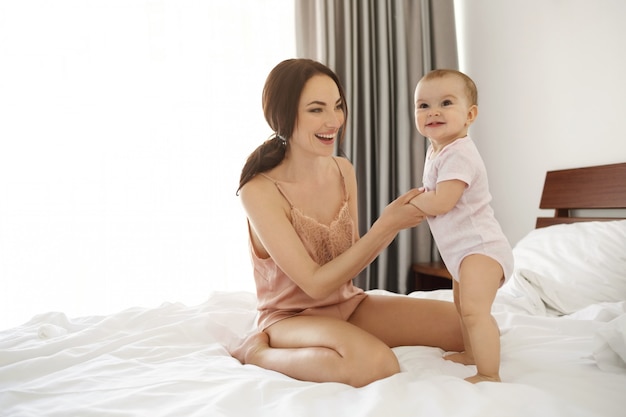 Mãe feliz jovem alegre em roupa de noite e sua filha bebê sorrindo jogando sentado na cama de manhã.