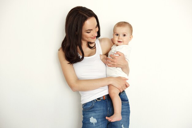 Mãe feliz nova que sorri guardando olhando sua filha do bebê sobre a parede branca.