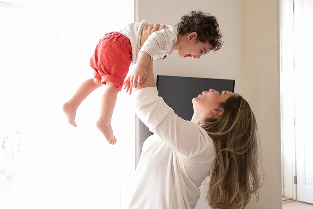 Mãe feliz segurando bebê animado nos braços, levantando a criança no ar. Vista lateral. Conceito de paternidade e infância