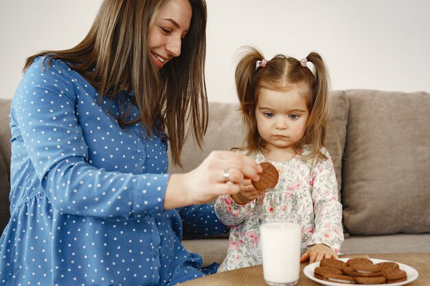 Mãe grávida em um vestido. A menina bebe leite. Mãe e filha gostam de biscoitos.