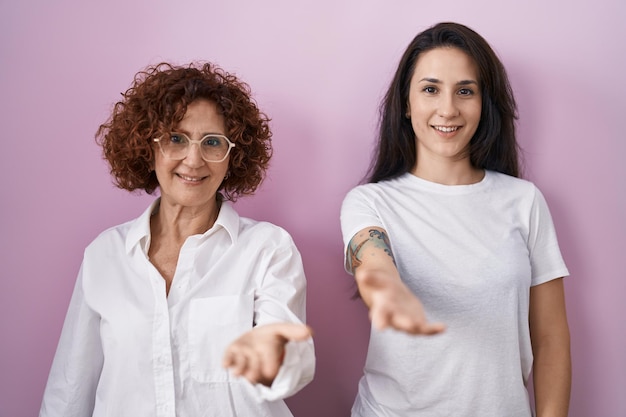 Mãe hispânica e filha vestindo camiseta branca casual sobre fundo rosa sorrindo alegremente oferecendo palma dando assistência e aceitação.