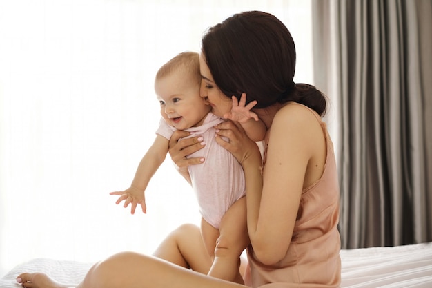 Mãe nova na roupa de noite que sorri abraçando beijando seu bebê que senta-se na cama sobre a janela. Olhos fechados.