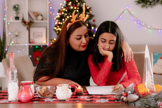 mãe satisfeita com bandana de rena olha para a carta que sua filha está escrevendo, sentada à mesa, aproveitando o Natal em casa