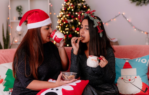 Mãe satisfeita com chapéu de Papai Noel bebe do copo e olha para a filha com coroa de azevinho comendo biscoito, sentada no sofá, aproveitando o Natal em casa