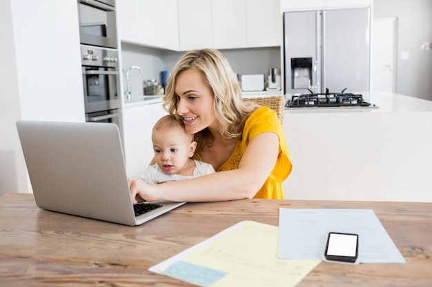 Mãe usando laptop com o bebé na cozinha