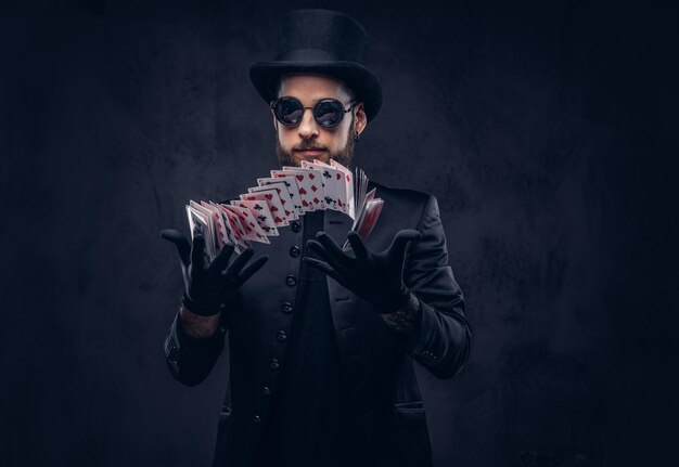 Mágico de terno preto, óculos escuros e cartola, mostrando truque com cartas de baralho em um fundo escuro.