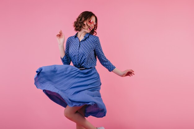 Magnífica modelo feminina em saia midi dançando e rindo na parede rosa. mulher caucasiana animada com roupas azuis, expressando emoções positivas.